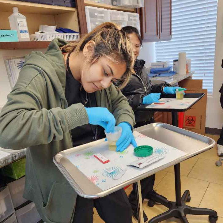 Medical assistant in scrubs preparing clinical equipment in an exam room.